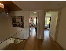 Interno di una casa con corridoio in parquet e pareti bianche, illuminato da luce naturale, con una scala con ringhiera in vetro e legno sulla sinistra. In fondo si aprono porte verso altre stanze, con una camera visibile a destra.