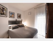 Camera da letto luminosa con letto matrimoniale e testiera nera in metallo, biancheria grigia e tre fotografie incorniciate sopra il letto. A destra una grande portafinestra con tende bianche lascia entrare luce naturale, accanto a un armadio in legno scuro.