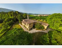 Veduta aerea di un complesso storico in pietra con tetti in coppi, torre campanaria e cortile interno, circondato da alberi e vialetti. Sullo sfondo si vedono campi coltivati, un centro abitato e montagne all’orizzonte.