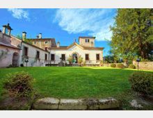 Cortile esterno di una villa storica con edifici intonacati chiari e dettagli in pietra, ingresso ad arco centrale e tetti con comignoli. In primo piano si vede un ampio prato verde con cespugli e lampioni, delimitato da un muretto e alberi sul lato destro.