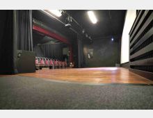 Interno di una piccola sala teatrale con palco in legno, pareti e tendaggi neri e luci di scena a soffitto. A sinistra sono visibili file di sedie rosse per il pubblico e una struttura ad arco, mentre a destra si nota una grande superficie chiara tipo schermo.