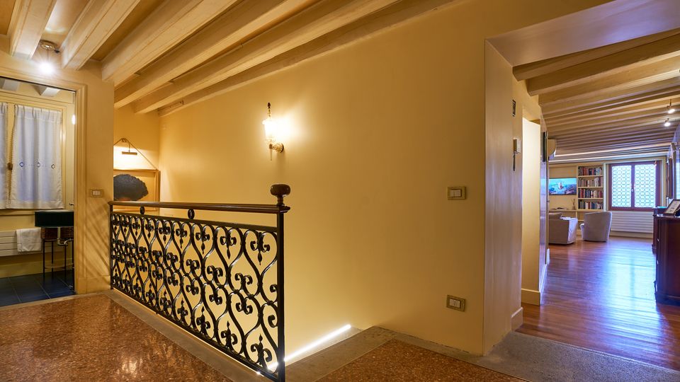 Interno di una casa con soffitto a travi in legno e pareti color crema, con corrimano in ferro battuto lungo un piccolo ballatoio su pavimento in terrazzo. A destra si apre un soggiorno con pavimento in legno, libreria e grande finestra, mentre a sinistra si intravede un bagno con finestra e tende bianche.