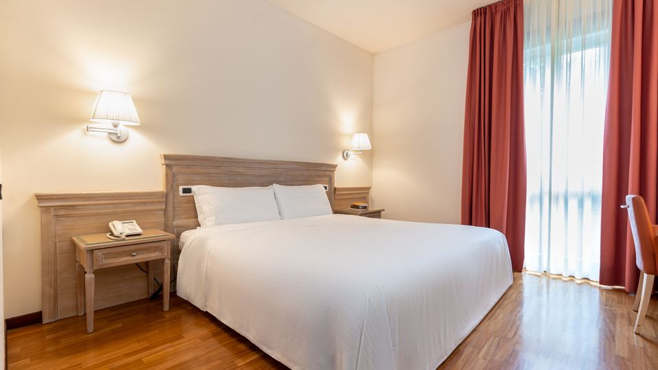 Camera da letto d’albergo con letto matrimoniale, testiera e comodini in legno e applique a parete. Pavimento in parquet e grande finestra a tutta altezza con tende rosse e velo bianco che lascia entrare luce naturale.