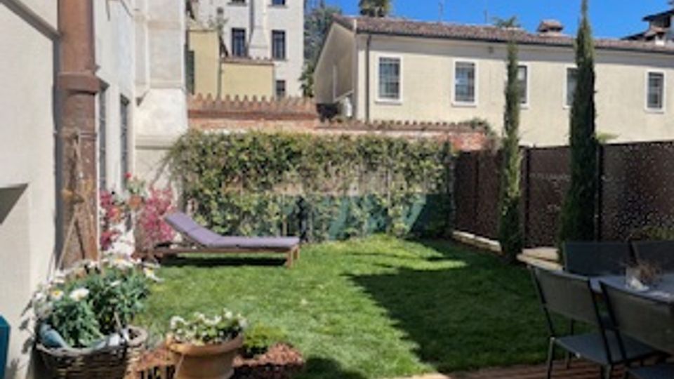 Piccolo giardino privato con prato curato, alcune piante in vaso e fioriere lungo il muro, e una sedia a sdraio appoggiata vicino alla recinzione. Sullo sfondo si vedono edifici residenziali a più piani e un camminamento in legno con tavolo e sedie sul lato destro.