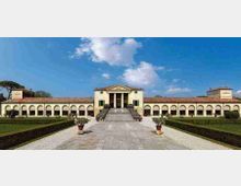 Facciata di una villa storica con corpo centrale neoclassico, portico a colonne e lunghe ali laterali ad arcate. Davanti si estende un viale in pietra con gradinata centrale, siepi geometriche e aiuole di giardino sotto un cielo azzurro.