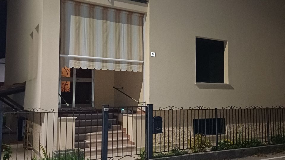 Facciata di una palazzina residenziale chiara vista di notte, con ingresso rialzato raggiungibile da una scalinata e una tenda a righe sopra la porta. Al piano superiore ci sono ampie finestre con balcone in ferro, e davanti all’edificio un cancello e una recinzione metallica lungo il marciapiede e la strada.