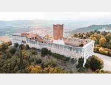 Veduta aerea di una grande fortezza in pietra su una collina, con una lunga cinta muraria e una torre quadrata centrale. Intorno si vedono alberi e un paesaggio collinare con strade e insediamenti in lontananza.