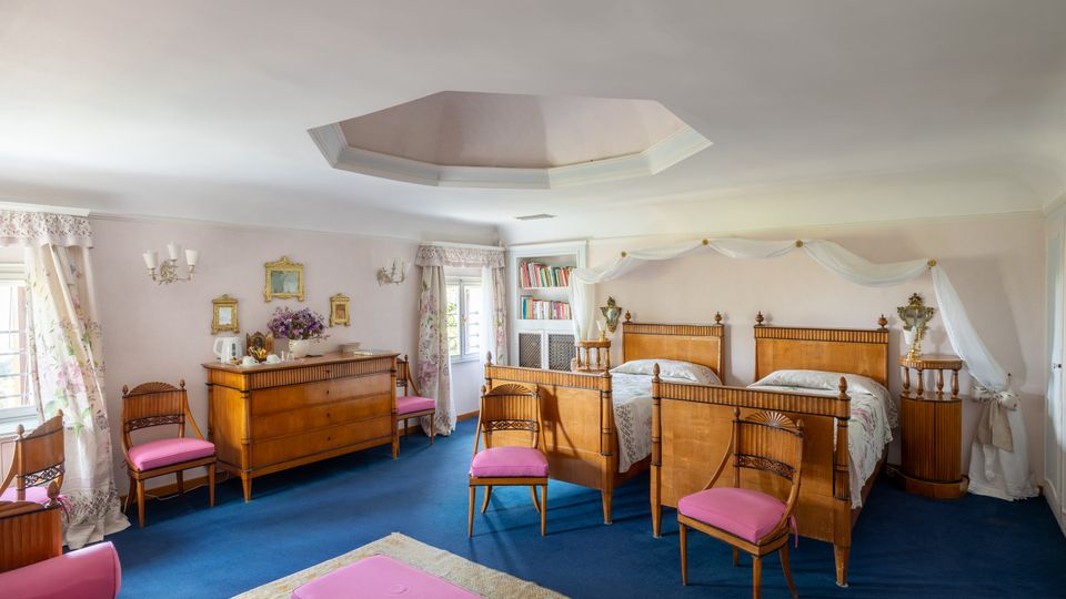 Ampia camera da letto in stile classico con moquette blu, due letti singoli in legno con drappeggi bianchi e comodini coordinati. La stanza è illuminata da finestre con tende floreali e arredata con comò, sedie imbottite rosa e mensole con libri.