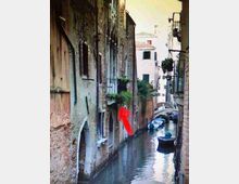 Stretto canale urbano tra alti edifici in muratura con finestre e balconi, con acqua che scorre lungo il passaggio. In fondo si vede un piccolo ponte ad arco e alcune barche ormeggiate lungo il canale.