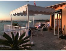 Terrazza panoramica sul mare al tramonto con pavimentazione in piastrelle, ringhiera bianca e una pergola con tenda ondulata. Sono presenti un tavolino con sedie, un lettino e piante in vaso, accanto alla facciata di un edificio con grandi vetrate.