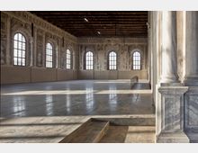 Ampia sala storica con pavimento in pietra lucida, soffitto a travi in legno e grandi finestre ad arco che illuminano pareti affrescate. In primo piano si vede un breve gradino e, sul lato destro, colonne in pietra e un piccolo corrimano metallico.