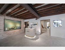 Interno di una galleria d’arte moderna con pavimento in cemento, pareti bianche e una parete in mattoni a vista con grande opera esposta. Al centro c’è un banco reception curvo illuminato, mentre il soffitto presenta travi in legno e tubazioni a vista con ingressi verso altre sale.