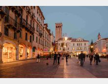 Piazza urbana al crepuscolo con ampia pavimentazione in pietra, edifici storici con portici e balconi sul lato sinistro e facciate chiare sullo sfondo. Al centro si nota una torre merlata e lampioni accesi, con negozi e locali illuminati lungo i portici.