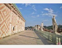 Terrazza panoramica in pietra lungo il fianco di un edificio storico con facciata decorata a motivi geometrici e finestre circolari. Sul lato esterno corre una balaustra con statue classiche e vista sui tetti della città sotto un cielo azzurro.