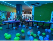 Interno di una sala tipo foyer/corridoio con pareti verdi e illuminazione a neon blu sul soffitto, con tavoli allestiti e numerosi palloncini sparsi sul pavimento. Sullo sfondo sono visibili grandi poster cinematografici e pannelli pubblicitari lungo le pareti.