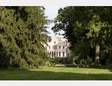 Vista di una villa storica in stile neogotico, con facciata chiara, archi e finestre alte, parzialmente nascosta tra grandi alberi. In primo piano si vede un ampio prato verde che conduce verso l’edificio immerso in un parco.