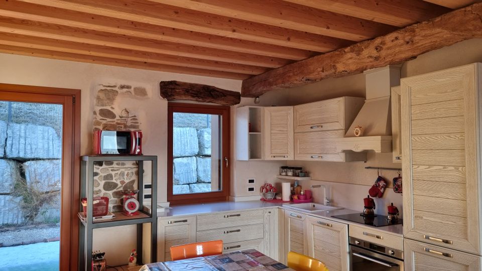 Cucina rustica con soffitto a travi di legno a vista e grande trave scura, mobili chiari a L con cappa e piano cottura. Due finestre e una porta finestra con cornici in legno affacciano su un muro in pietra; a sinistra una nicchia in pietra e una mensola metallica con piccoli elettrodomestici.