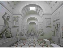 Interno di una galleria museale neoclassica con volte a botte e archi, illuminata da un lucernario centrale. Lungo le pareti sono esposti bassorilievi e statue in marmo su piedistalli, con pavimento a piastrelle geometriche bianche e nere e panche basse.