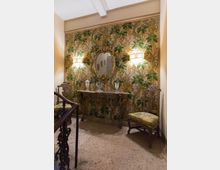 Piccolo pianerottolo interno con carta da parati floreale verde, console antica in legno e specchio ovale dorato al centro, illuminati da due applique a parete. Ai lati ci sono due sedie imbottite e in primo piano si vede la ringhiera della scala con pavimento in pietra.