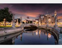 Veduta al crepuscolo di un canale urbano con un ponte in pietra ad arco decorato da statue, che si riflette nell’acqua calma. Sullo sfondo si vede una grande basilica con cupole e campanile, circondata da lampioni e edifici lungo le rive.