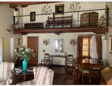 Interno di un soggiorno con soppalco in legno e ringhiera metallica, soffitto con travi a vista e arredi classici in legno. Nella zona inferiore ci sono poltrone e tavolino, una zona pranzo vicino a una finestra con tende, mentre sul soppalco sono presenti una panca e mobili antichi con quadri e applique alle pareti.