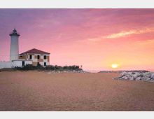 Faro bianco con edificio annesso su una spiaggia sabbiosa, affacciato sul mare. All’orizzonte si vede il sole al tramonto con cielo rosa e una scogliera di massi sulla destra.