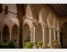 Chiostro in mattoni con porticato ad archi e colonne in pietra con capitelli scolpiti, sotto un soffitto a cassettoni in legno. Al centro si vede un piccolo giardino con piante e vasi disposti lungo il camminamento.