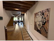 Interno di un’abitazione moderna con lungo corridoio in parquet, soffitto con travi in legno a vista e pareti bianche. A sinistra si vede una scala in legno e in fondo una zona giorno luminosa con grandi finestre; sulla parete destra è appeso un grande quadro in stile pop art.