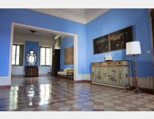 Interno di un'elegante sala con pareti azzurre, pavimento in marmo con motivo a scacchiera e un mobile antico decorato con pannelli dipinti. Sono presenti un grande specchio ornato, quadri appesi alla parete e una lampada da terra accanto al mobile.