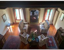 Soggiorno con pavimento in legno e travi a vista, arredato con due divani e quattro poltrone disposti attorno a un tavolino centrale su tappeti persiani. Due grandi finestre con tende e un grande specchio decorato tra di esse illuminano la stanza, con radiatore e mobili lungo le pareti.