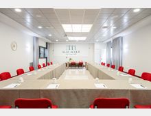 Sala conferenze moderna con tavoli disposti a U coperti da tovaglie beige, sedie rosse e blocchi per appunti. Soffitto a pannelli con illuminazione incassata, pareti bianche con tende grigie e parete frontale con insegna “Alle Acque”.