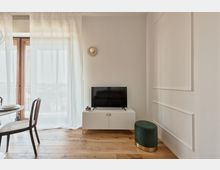 Interno di un soggiorno moderno e luminoso con pavimento in legno, pareti bianche con boiserie e una grande portafinestra con tende leggere. Un mobile TV bianco con televisore e piccola pianta è affiancato da un pouf verde, con tavolo da pranzo e sedie visibili sul lato sinistro.