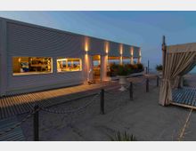 Edificio moderno sul lungomare con facciata a pannelli orizzontali e ampie finestre illuminate che mostrano l’interno di un bar o ristorante. In primo piano c’è una passerella in legno sulla sabbia con corde e paletti, vasi con piante e una struttura tipo gazebo con tende, con il mare sullo sfondo al crepuscolo.