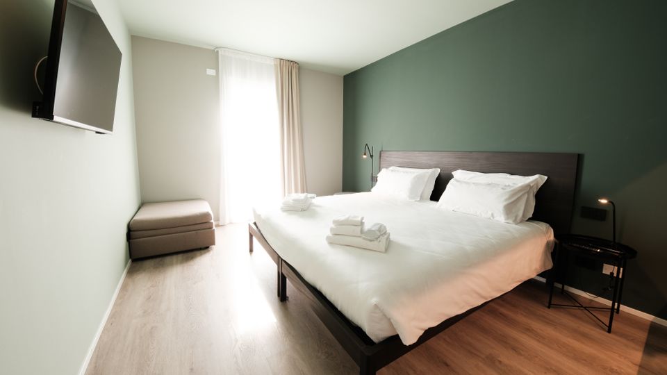 Camera da letto moderna con letto matrimoniale in lenzuola bianche e testiera scura, parete verde e pavimento in legno chiaro. Di fronte al letto ci sono una TV a parete e una panca imbottita, con una grande finestra a tutta altezza con tende che illumina l’ambiente.