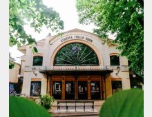Facciata del “Cinema Italia Eden” con grande arco centrale vetrato a raggiera e pensilina in ferro battuto sopra l’ingresso. L’edificio è circondato da alberi e vegetazione, con una panchina e un vialetto davanti.