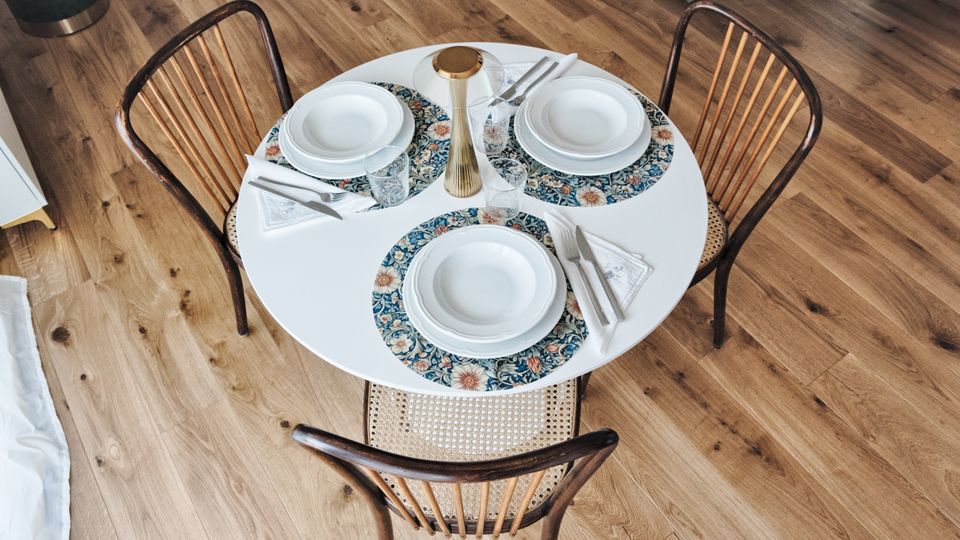 Tavolo da pranzo rotondo bianco apparecchiato per tre con piatti, posate, bicchieri e tovagliette floreali, visto dall’alto. Intorno ci sono tre sedie in legno con seduta intrecciata su un pavimento in parquet chiaro.