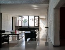 Interno di un ampio soggiorno moderno con pavimento lucido, pareti in cemento grezzo e divani neri disposti attorno a un tavolino basso. Grandi finestre a tutta parete illuminano lo spazio e mostrano l’esterno con un muro bianco decorato da piccoli elementi geometrici.