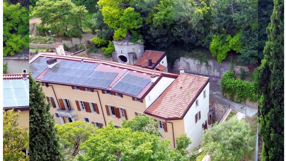 Veduta dall’alto di un edificio residenziale color crema con persiane in legno, tetto in coppi e ampi pannelli solari. La casa è circondata da alberi fitti e vegetazione, con un piccolo cortile e un vialetto lungo un muro di contenimento sul lato.