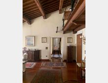 Interno di un’abitazione con soffitto alto in legno a travi a vista e soppalco con ringhiera metallica. Pavimento in parquet con tappeti, mobili in legno e quadri alle pareti, con un corridoio centrale che conduce ad altre stanze.