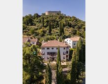 Veduta di un borgo collinare con una villa a più piani dai muri chiari e tetto in coppi, circondata da cipressi e altre case. Sullo sfondo, una collina ricoperta di alberi e ulivi culmina in una fortezza in pietra.