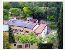 Veduta dall’alto di un edificio residenziale color crema con persiane in legno, tetto in coppi e ampi pannelli solari. La casa è circondata da alberi fitti e vegetazione, con un piccolo cortile e un vialetto lungo un muro di contenimento sul lato.