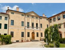 Facciata di una villa storica a due piani con intonaco chiaro, frontone centrale e quattro lesene che incorniciano un balconcino sopra un portale in legno. Davanti si vede un vialetto di ghiaia con vasi di piante e un pergolato con rampicanti nel cortile, tra edifici adiacenti.