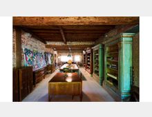Interno rustico con soffitto a travi in legno e pareti in mattoni a vista, arredato come studio o biblioteca con grandi scaffalature e armadi antichi. Al centro ci sono tavoli in legno e lampade, con quadri colorati appoggiati lungo la parete sinistra e una finestra luminosa sul fondo.