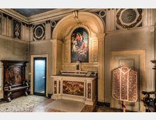 Interno di una piccola cappella con altare in marmo sotto un arco e grande dipinto religioso sopra, circondata da pareti decorate con stucchi e medaglioni sul soffitto. A lato si vedono panche e arredi in legno, una porta vetrata e un paramento liturgico esposto in una nicchia.