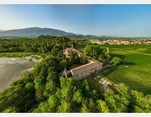 Veduta aerea di un complesso rurale con edifici in muratura e tetti in coppi, immerso in un’area boscata tra campi coltivati e una zona umida. Sullo sfondo si estendono una pianura con piccoli centri abitati e una catena montuosa sotto un cielo sereno.