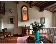 Soggiorno in stile rustico con travi in legno a vista, pavimento in legno e pareti chiare con quadri e una nicchia decorativa illuminata. Arredi classici con tavolo e sedie, cassettiera, divano a righe e tavolino rotondo con vaso turchese e composizione floreale su tappeto.