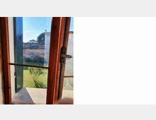 Finestra in legno con ante aperte e chiusura metallica, vista dall’interno. Attraverso i vetri si vede un prato verde con un piccolo albero e, sullo sfondo, un muro in pietra e alcuni edifici sotto un cielo sereno.