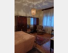 Camera da letto con arredi classici: letto con struttura in ottone, grande armadio in legno scuro con ante a specchio e lampadario a gocce sospeso. Accanto a una finestra con tende leggere c’è una poltrona imbottita, con tappeti sovrapposti su pavimento in parquet.