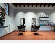 Interno di un salone da parrucchiere con pavimento in cotto e soffitto con travi in legno a vista, dotato di due postazioni con specchi e sedie nere. Sulle pareti sono presenti mensole e scaffali con prodotti per capelli e una finestra laterale con grata.