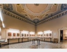 Sala espositiva in un edificio storico con soffitto alto decorato da stucchi e affreschi e pavimento in terrazzo. Lungo le pareti e su tavoli centrali sono esposte piccole opere e pannelli, illuminate da luci lineari a parete.